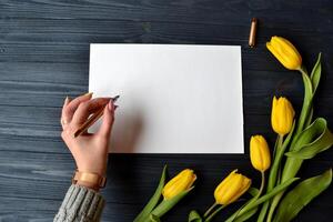 donna è scrittura un' Messaggio su il bianca carta. primavera ancora vita. giallo tulipani, bianca carta su il blu Vintage ▾ scrivania. penna nel femmina mano. foto