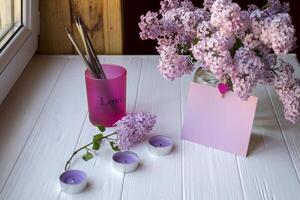 un' bicchiere con matite, lilla nel vaso, vuoto rosa carta e decorativo elementi su un' bianca di legno tavolo. foto