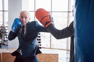 un uomo d'affari calvo arrabbiato batte una pera da boxe in palestra. concetto di gestione della rabbia foto