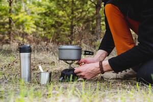 attrezzatura per un' escursione, metallo utensili per mangiare, un' turista prepara prima colazione nel natura, impostato fuoco per un' gas bruciatore, Bollire acqua, un' tazza, un' thermos con tè. foto