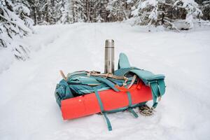 escursioni a piedi attrezzatura. un' luminosa escursioni a piedi zaino bugie su il neve.. thermos con caldo bevanda bugie su zaino. escursioni a piedi nel inverno. sopravvivenza nel foreste foto