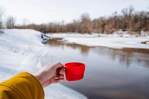 un' rosso boccale nel un' di persona mano, un' mano Tenere un' bicchiere contro il sfondo di il fiume, un' turista potabile tè nel natura, un' viaggiatore inverno escursione, primavera fusione fiumi foto