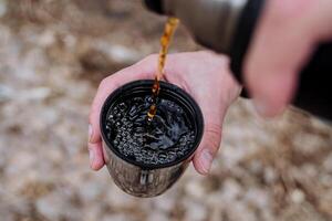 un' bicchiere di tè è tiro avvicinamento, versare tè in un' boccale su superiore, un' caldo Marrone bere, un' termo tazza, un' thermos versa acqua, un' mano detiene un' tazza di caffè. foto