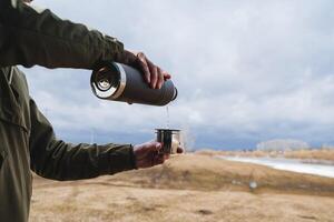 versare tè a partire dal un' thermos in un' boccale contro un' nuvoloso cielo, un' turista fuoriuscite un' caldo bevanda in il suo bicchiere, il concetto di un' vuoto bottiglia nel campeggio condizioni. foto