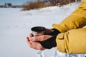 mani hold un' boccale di caldo tè contro il sfondo di neve nel inverno. giallo manica di giù giacca. caldo il tuo mani con caldo tè. foto
