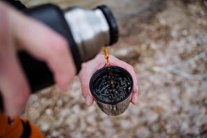 un' thermos versare tè in un' tazza, un' mano detiene un' bicchiere di caldo tè su un' escursione, bolle nel un' tazza, caldo bevande nel freddo tempo atmosferico, turista utensili, caffè nel il mattina. foto