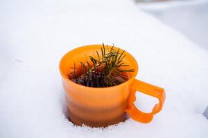 un arancia plastica boccale con un' abete rosso ramo bugie nel il neve. luminosa piatti contro il sfondo di puro neve. aroma a partire dal abete rosso. inverno minimalista tiro foto