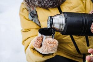 thermos mano avvicinamento. versare il tè in il temico cerchio. giallo giacca. un' mano detiene un' bicchiere di caffè. nero metallo nave, acciaio bottiglia. foto