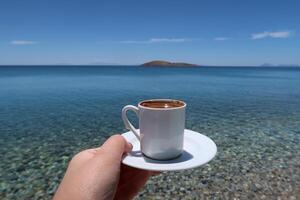 tradizionale Turco caffè tazza con modelli. donna mano Tenere il tazza su un' soleggiato estate giorno, esotico mare sfondo foto