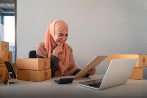 musulmano donne vendita in linea a casa con scatola. vendita in linea con scatola per accettare ordine a partire dal cliente. pmi attività commerciale idea. pacco consegna. musulmano donna Lavorando smartphone e il computer portatile a casa foto