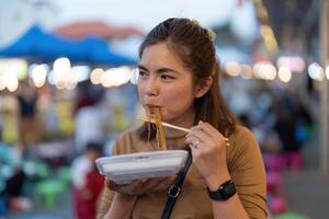 giovane asiatico turista donna viaggiatore mangiare tampone tailandese spaghetto a notte mercato, tradizionale chiang Mai Tailandia strada cibo. foto