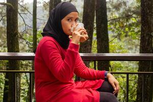 un asiatico musulmano donna nel rosso lungo manica camicia potabile un' plastica tazza di ghiaccio caffè su il balcone con natura o alberi sfondo foto