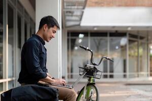 asiatico uomo d'affari con bicicletta utilizzando il computer portatile e seduta al di fuori il ufficio costruzione. uomo pendolarismo su bicicletta partire per opera. eco amichevole veicolo, sostenibile stile di vita concetto foto