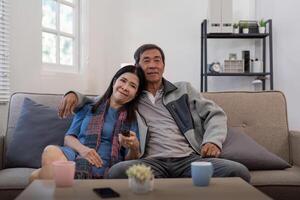 anziano coppia Guardando tv e divano nel rilassare per film o serie nel vivente camera a casa. anziano uomo e donna con caffè e a distanza insieme per mutevole canale e in linea divertimento foto