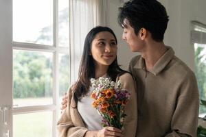 romantico giovane asiatico coppia Abbracciare una persona con Tenere fiori e sorridente nel vivente camera a casa. autunno nel amore. San Valentino concetto foto