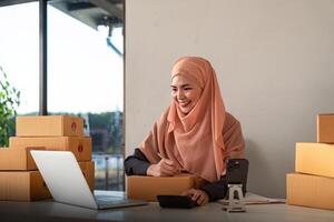 musulmano donne vendita in linea a casa con scatola. vendita in linea con scatola per accettare ordine a partire dal cliente. pmi attività commerciale idea. pacco consegna. musulmano donna Lavorando smartphone e il computer portatile a casa foto