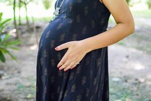 un' incinta indiano signora pose per all'aperto gravidanza sparare e mani su gonfiarsi, indiano incinta donna mette sua mano su sua stomaco con un' maternità vestito a società parco, incinta al di fuori maternità sparare foto