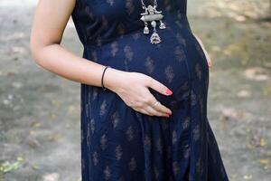 un' incinta indiano signora pose per all'aperto gravidanza sparare e mani su gonfiarsi, indiano incinta donna mette sua mano su sua stomaco con un' maternità vestito a società parco, incinta al di fuori maternità sparare foto