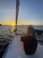 un' giovane ragazzo si siede su il arco di un' barca a vela a tramonto foto