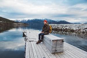 uomo viaggiatore con uno zaino giallo che indossa un cappello rosso seduto sul molo di legno sullo sfondo della montagna e del lago. spazio per il tuo messaggio di testo o contenuto promozionale foto