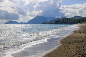 bellissimo spiaggia scena con montagna, morbido sabbia, onde, e nuvoloso cielo impostato contro luminosa luce del sole foto