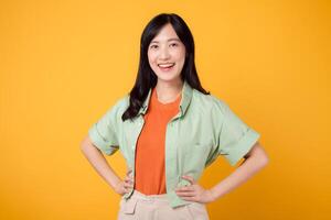 radiante felicità giovane asiatico donna 30s indossare un' verde camicia su un arancia camicia vetrine gioioso viso mentre in piedi con akimbo braccia. puro gioia con vivace Immagine su un' giallo sfondo. foto