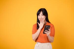 curioso asiatico donna nel sua anni '30, indossare arancia camicia, utilizzando smartphone con cazzotto su mano cartello su vivace giallo sfondo. svelare nuovo App meraviglia. foto