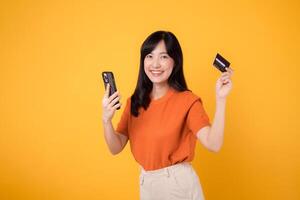 energico giovane asiatico donna nel sua anni '30, utilizzando smartphone e Tenere credito carta su giallo sfondo. veloce in linea acquisti. foto