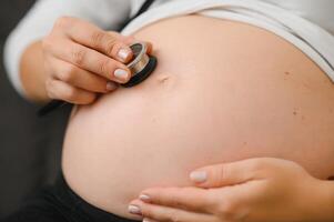 il madre è utilizzando stetoscopio il medico ascolta per il suono di il bambino nel il stomaco. asiatico incinta donna utilizzando stetoscopio ascoltando sua bambino su divano. foto