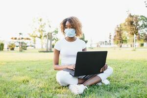 africano americano ragazza nel un' protettivo maschera Lavorando su un' il computer portatile nel il parco su il erba. il concetto di a distanza opera. foto