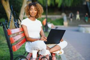 allegro giovane nero ragazza su un' di legno panchina nel un' parco è utilizzando il il computer portatile abbagliante sorridente brasiliano femmina all'aperto su il panchina con il netbook nel mani e piccolo pelle Borsa su il sinistra foto