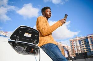 elettrico macchine, ev concetto, eco amichevole carburante. ritratto di giovane sorridente nero uomo, ricarica il suo moderno lusso elettrico auto foto