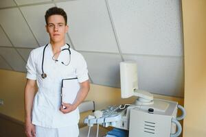 affascinante giovane maschio medico , seduta a il suo ufficio vicino ultrasuono lettura macchina. bello amichevole medico godendo Lavorando a il Ospedale, copia spazio. ultrasonico concetto foto