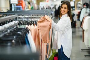 saldi, Abiti , acquisti, moda e persone concetto - contento giovane donna la scelta fra Due camicie nel centro commerciale o capi di abbigliamento memorizzare foto