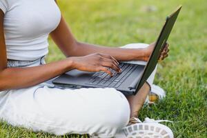 giovane sorridente nero donna seduta all'aperto su erba con computer portatile, digitando, fare surf Internet. tecnologia, comunicazione, formazione scolastica e a distanza Lavorando concetto, copia spazio foto