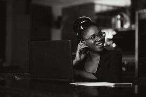 bellissimo afro americano ragazza nel casuale Abiti è utilizzando un' il computer portatile e sorridente mentre seduta nel bar foto