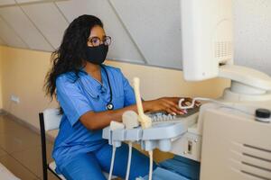 giovane femmina afroamericano medico Lavorando su moderno ultrasuono attrezzatura. operatore di ultrasuono lettura macchina seduta e guardare a il tenere sotto controllo, in attesa per paziente. foto