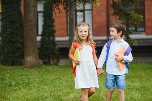 contento bambini - ragazzo e ragazza con libri e zaini su il primo scuola giorno. eccitato per essere indietro per scuola dopo vacanza. pieno lunghezza all'aperto ritratto. foto