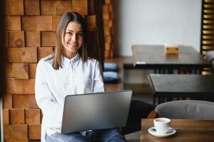 ritratto di un' giovane femmina libero professionista utilizzando il computer portatile computer per distanza lavoro mentre seduta nel moderno caffè negozio interno, inteligente bionda donna Lavorando su netbook durante mattina prima colazione nel bar bar foto