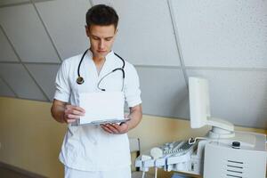 affascinante giovane maschio medico , seduta a il suo ufficio vicino ultrasuono lettura macchina. bello amichevole medico godendo Lavorando a il Ospedale, copia spazio. ultrasonico concetto foto