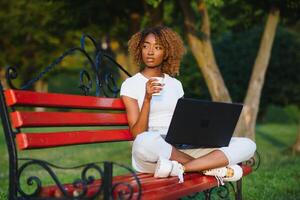 bellissimo sorridente giovane africano donna seduta su un' panchina all'aperto, Lavorando su il computer portatile computer foto