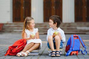 primario formazione scolastica, amicizia, infanzia e persone concetto - gruppo di contento elementare scuola studenti con zaini. Due scolari seduta su libri vicino scuola e mangiare un Mela. foto