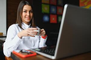 ritratto di un' giovane femmina libero professionista utilizzando il computer portatile computer per distanza lavoro mentre seduta nel moderno caffè negozio interno, inteligente bionda donna Lavorando su netbook durante mattina prima colazione nel bar bar foto