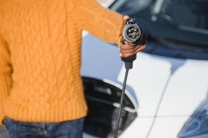 vicino su di maschio mano Tenere elettrico ricarica spina per lusso rosso macchina. africano uomo d'affari ricarica il suo auto su stazione. eco amichevole trasporto. foto