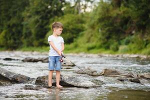 carino ragazzo nel bianca t camicia pesca nel il fiume e ha divertimento, sorrisi. vacanza con bambini, vacanze, attivo fine settimana concetto foto