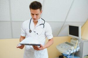 affascinante giovane maschio medico , seduta a il suo ufficio vicino ultrasuono lettura macchina. bello amichevole medico godendo Lavorando a il Ospedale, copia spazio. ultrasonico concetto foto