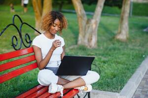 bellissimo sorridente giovane africano donna seduta su un' panchina all'aperto, Lavorando su il computer portatile computer foto