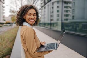 sorridente femmina libero professionista Lavorando il computer portatile mentre in piedi al di fuori su sfondo di ufficio edificio foto