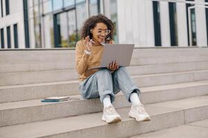 femmina libero professionista avere un' video conferenza con collega e agitando Ciao seduta su le scale al di fuori foto