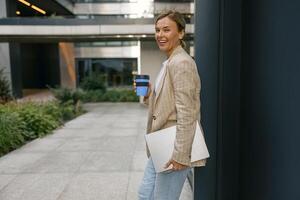 sorridente attività commerciale donna in piedi con il computer portatile e prendere lontano caffè su elegante ufficio cortile foto